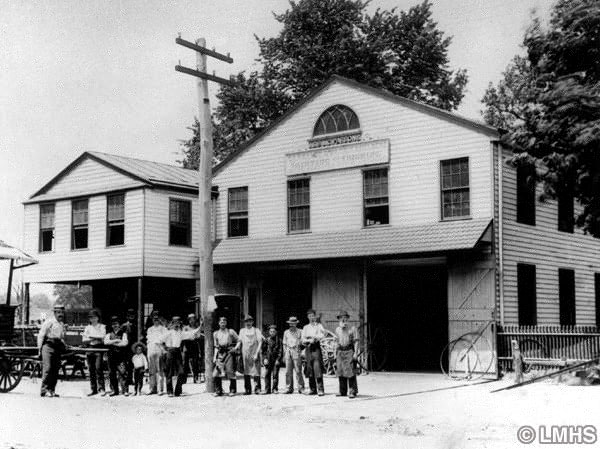 row of men and boys stand in front of 2 buildings with garage-size doors