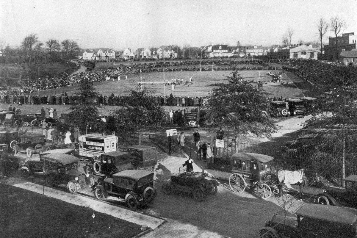 cars in front, fans crowd around, teams ion  football field in background