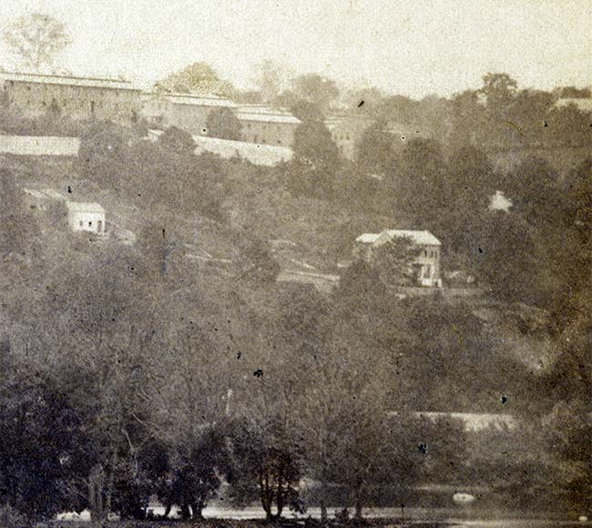 wooded hillside with large 2-story buildings at top, smaller buildings in middle, river at bottom