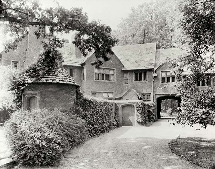 driveway leads from a portcullis beside a round gatehouse with a pointed slate-tiled roof and ivy-covered walls to an ornate mansion