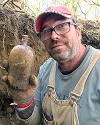 man in overalls stands in deep pit holding a dirt-caked glass bottle