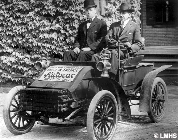 two men wearing suits sit in the single seat of a small early vehicle with a shaft in place of a steering wheel