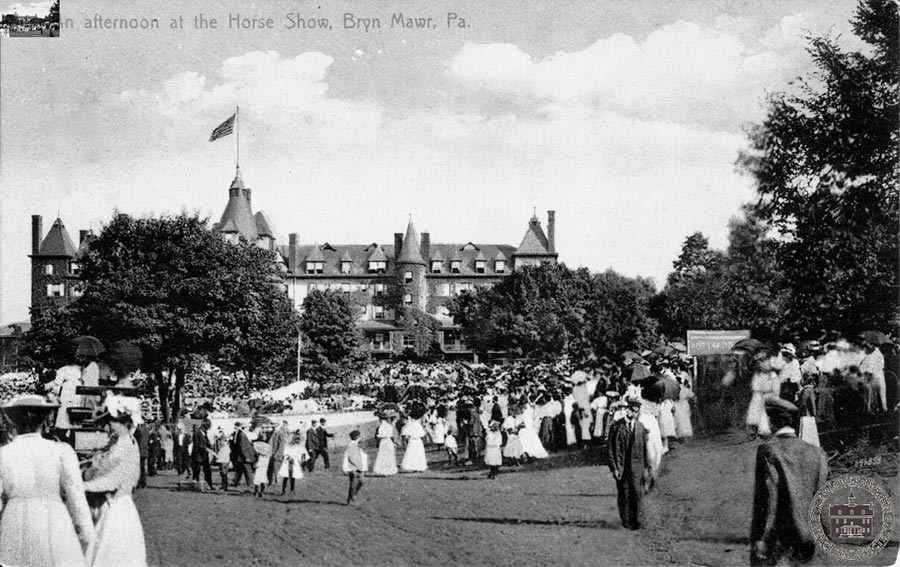 A well-dressed crowd, ladies in white, mingles on the grounds in front of the hotel