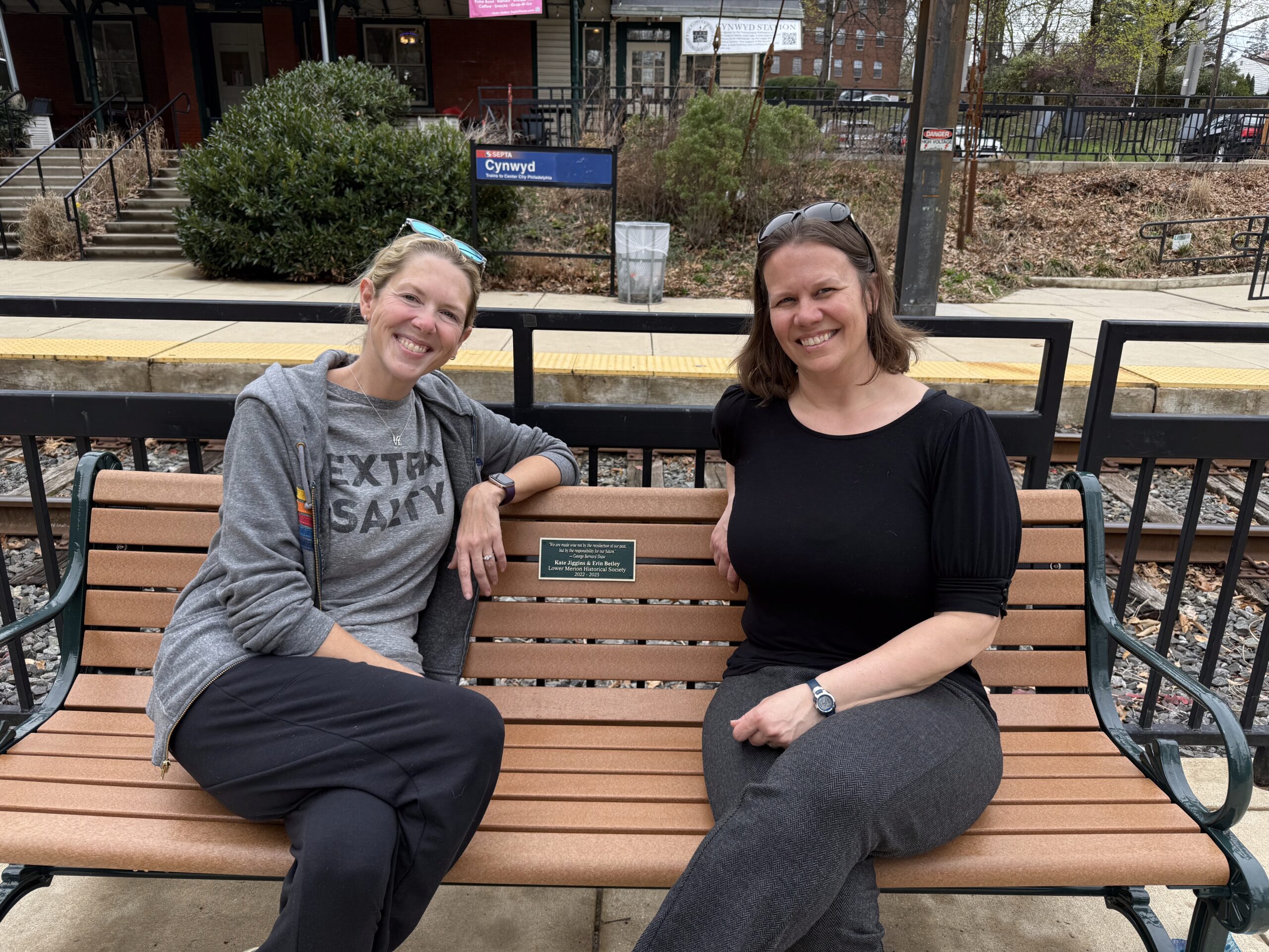 Former Society President Kate Jiggins and former Vice President Erin Betley sit on a commemorative bench at the Cynwyd Station.
